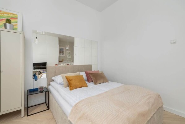 A bright bedroom with a double bed in beige bedding, a mirror above, and white walls for an airy look.
