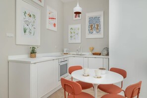 A modern white kitchenette with a circular dining table and orange chairs. Colorful artwork decorates the walls.
