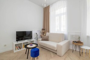 A cozy living area with a light sofa, TV, and a round blue coffee table. Large windows let in plenty of light.
