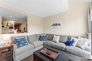 Living area with floor to ceiling windows, wood-burning fireplace, flat screen TV, hardwood flooring and cozy sectional which pulls out to offer an additional queen-sized bed.