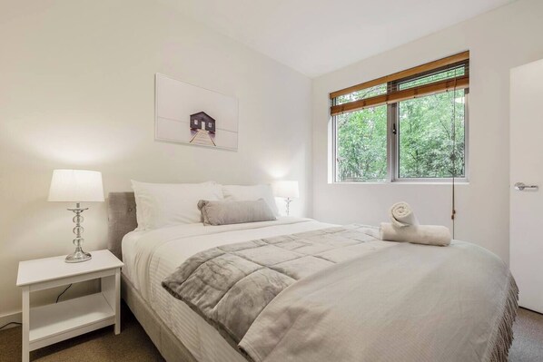 A bright bedroom has a queen bed dressed in linen, a built-in wardrobe and lots of natural light coming in through the window