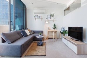 A living area is furnished with a comfortable L-shaped couch, small coffee table, plants and a flat-screen Smart TV