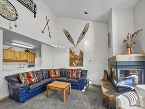 A living room with a blue sectional sofa, colorful throw pillows, and a wooden coffee table. There's a fireplace, wall art, and rustic decor like snowshoes and paddles mounted on the walls.