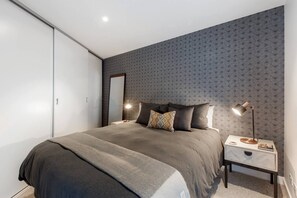A chic master bedroom features a plush queen bed dressed in hotel-quality linen, trendy bedside tables, a back feature wall and convenient built-in wardrobes