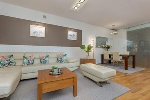 A bright living room with a beige sofa, ottoman, and a wooden coffee table leading to the dining area.