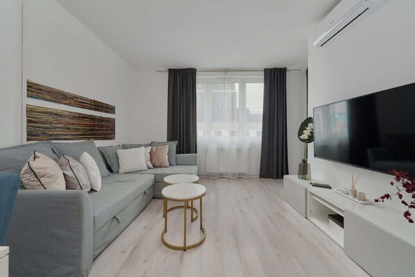 A bright living room with a light grey sofa, gold-accented coffee table, and wall-mounted TV.
