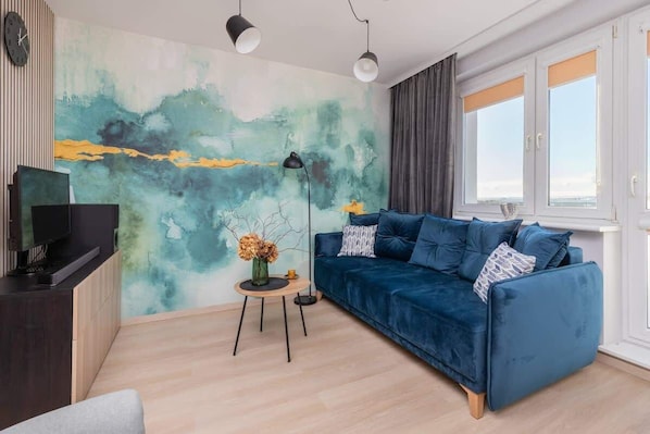 A bright living room featuring a teal watercolor-inspired accent wall, a plush navy sofa, and large windows that let in plenty of natural light.

