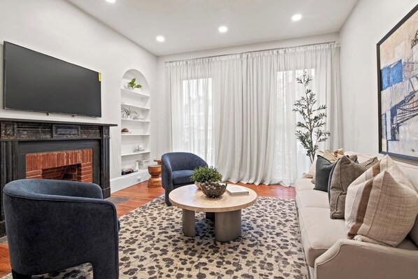 A cozy living room featuring a stylish setup with a large flat-screen TV mounted above a modern fireplace. A plush, inviting couch occupies the center, adorned with soft throw pillows. A sleek center table sits in front, adding a touch of elegance to