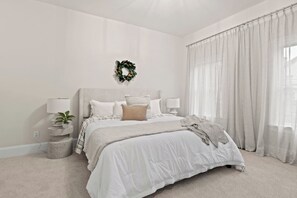 n this minimalist white bedroom, tranquility reigns with clean white linen dressing a neatly made bed. Airy curtains frame the windows, allowing soft natural light to fill the room. A simple yet stylish bedside table complements the serene atmosphere
