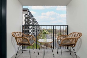 A cozy balcony setup with two rattan chairs and a small table, overlooking an urban view with blue skies and neighboring buildings.

