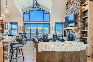 A spacious living room featuring a stone fireplace, large windows with a mountain view, a couch draped with a blanket, and a kitchen area with a counter and stools.