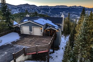 A mountain house with a snow-covered roof and wooden deck is surrounded by trees, with snow-capped mountains and a scenic valley visible in the background.