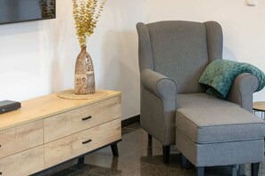 A close-up of a comfortable reading nook with a grey armchair, matching footstool, and a decorative vase on a wooden side table.

