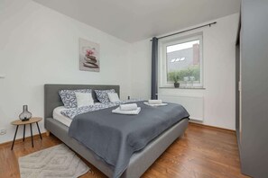 A cozy bedroom featuring a grey bedspread, wooden floors, and a simple nightstand with a neutral color scheme for a peaceful ambiance.

