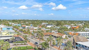 Aerial view of a suburban area with a mix of residential housing, commercial buildings, trees, and clear blue sky with scattered clouds.