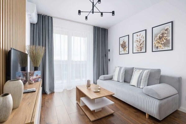 A bright living room featuring a grey sofa, wooden coffee table, and framed artwork on the wall, with floor-to-ceiling curtains allowing natural light to fill the space.

