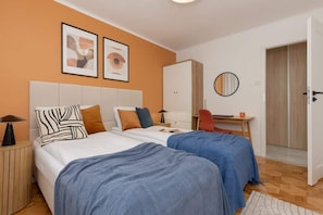A stylish bedroom with an orange accent wall, blue bedding, and framed art above the bed.