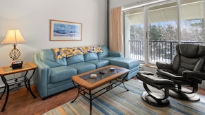 A cozy living room with a blue sofa, a patterned throw, a black recliner, a coffee table with items on it, a side table with a lamp, and a large window with a view of a balcony and trees.