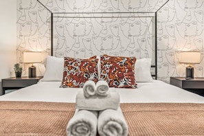 Immerse yourself in this stylish bedroom featuring bold, artistic wallpaper and plush pillows. You'll love the cozy ambiance created by the soft lighting and perfectly arranged decor. A perfect retreat to relax and unwind!