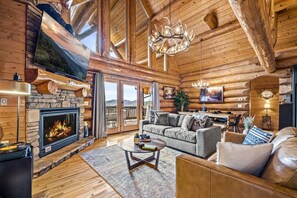 The expansive living room boasts high vaulted ceilings with log beams, a grand antler chandelier, and a stone fireplace, creating a warm and inviting atmosphere.