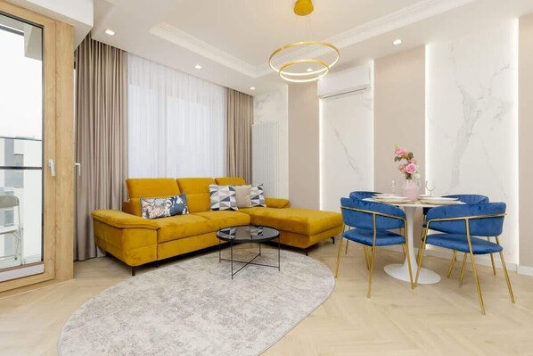 A modern living room with a yellow sofa, blue armchairs, and a minimalist table setup, featuring bright decor and ample lighting.