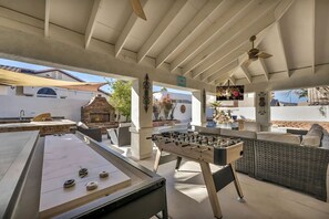 There's a shuffleboard table, foosball table, a seating area with a 75-inch TV, an outdoor kitchen space with a grill, and a fireplace, all under a covered patio. Looks like a fantastic spot for outdoor fun and relaxation!