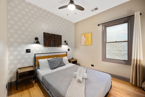 Cozy bedroom with a patterned wallpaper wall, a large mirror, a queen size bed with grey linens, a bedside table with a lamp and towels, and a window with white curtains.