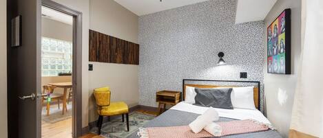 Cozy bedroom with a red zebra-patterned accent wall, a queen size bed with grey linens, a ceiling fan, a vanity area with a mirror and chair, and a window with white curtains.