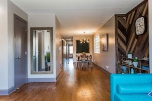 Wide view of the dining area with chairs, a glass block window, a modern chandelier, and a view into a living room with a fireplace and a leather armchair.