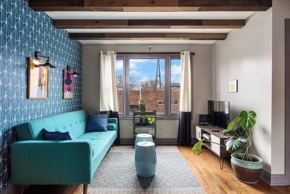 A close view living room with exposed wooden beams, a patterned wood accent wall, large windows, a leather sofa, a patterned rug, and several houseplants.