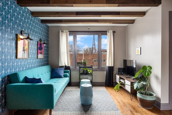 A close view living room with exposed wooden beams, a patterned wood accent wall, large windows, a leather sofa, a patterned rug, and several houseplants.
