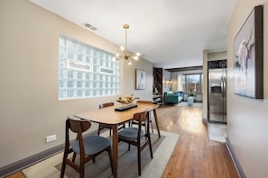 close view of the dining area with chairs, a glass block window, a modern chandelier, and a view into a living room with a fireplace and a leather armchair.