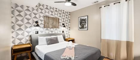 Cozy bedroom with a patterned wallpaper wall, a large mirror, a queen size bed with grey linens, a bedside table with a lamp and towels, and a window with white curtains.