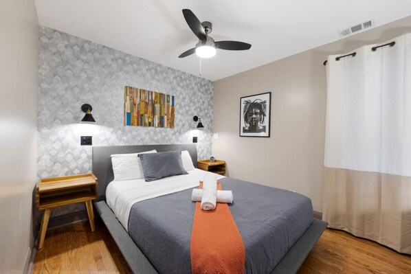 Cozy bedroom with a patterned wallpaper wall, a large mirror, a queen size bed with grey linens, a bedside table with a lamp and towels, and a window with white curtains.