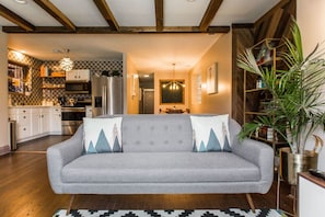 Close view of the living room with exposed wooden beams, a patterned wood accent wall, large windows, a leather sofa, a patterned rug, and several houseplants.