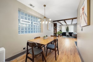 The dining area with chairs, a glass block window, a modern chandelier, and a view into a living room with a fireplace and a leather armchair.