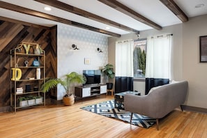 Back View of the living room with exposed wooden beams, a patterned wood accent wall, large windows, a leather sofa, a patterned rug, and several houseplants.