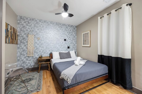 Cozy bedroom with a patterned wallpaper wall, a queen sized bed with grey linens, a ceiling fan, a vanity area with a mirror and chair,and a window with white curtains.