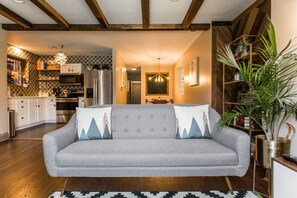Close view of the living room with exposed wooden beams, a patterned wood accent wall, large windows, a leather sofa, a patterned rug, and several houseplants.