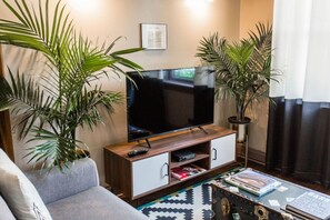 Close view of the living room with a grey sofa, a wooden TV stand, large potted palm plants, a patterned rug, and a collection of books on a vintage trunk.