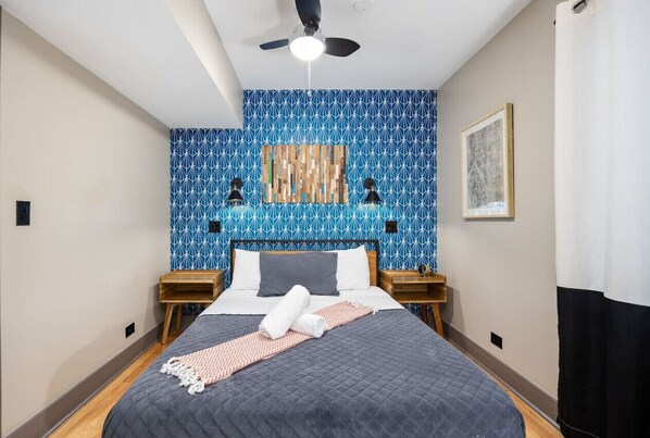 Cozy bedroom with a patterned wallpaper wall, a large mirror, a queen size bed with grey linens, a bedside table with a lamp and towels, and a window with white curtains.