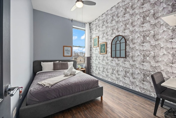 A simply furnished room with a plush armchair, a wooden table with and a framed painting on a blue ash paper wall.