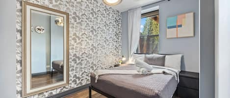 A cozy bedroom with a patterned wallpaper wall, a large mirror, a queen size bed with grey linens, a bedside table with a lamp and towels, and a window with white curtains.
