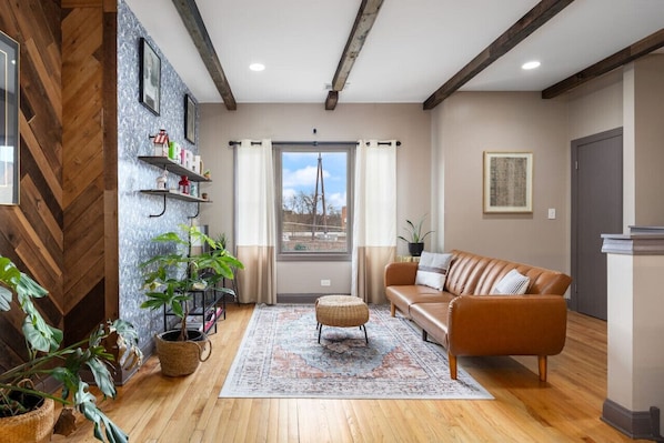 Cozy living room with exposed wooden beams, a patterned wood accent wall, large windows, a leather sofa, a patterned rug, and several houseplants.