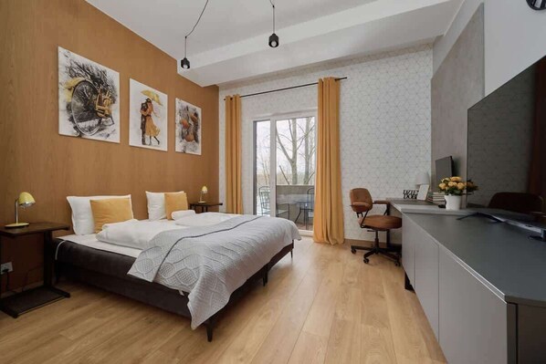 A spacious bedroom with a large bed, modern wall art, and a workspace by the window, illuminated by natural light.