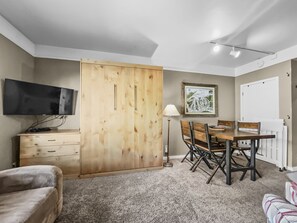 A cozy living room with a wall-mounted TV, a fold-out bed, a dining table with four chairs, a floor lamp, a cabinet, and a framed picture on the wall.