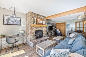 Living room with a stone fireplace, a mirror above the mantel, a blue sectional sofa, a wooden coffee table, a desk with a chair, and a TV near a wooden door.