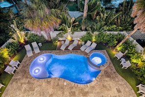 Twilight Private Heated Pool & Spa. The patio has chaise lounges and chairs surrounding the pool. Landscape lights illuminate the tropical gardens.