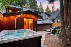 Pine trees surround the backyard, featuring an outdoor dining area, a BBQ grill, and a luxurious hot tub.
