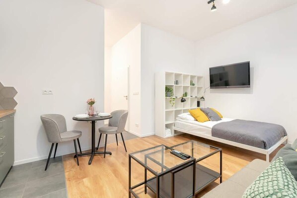 A cozy studio room with a grey sofa bed, yellow accent pillows, a small dining table, and a wall-mounted TV.
